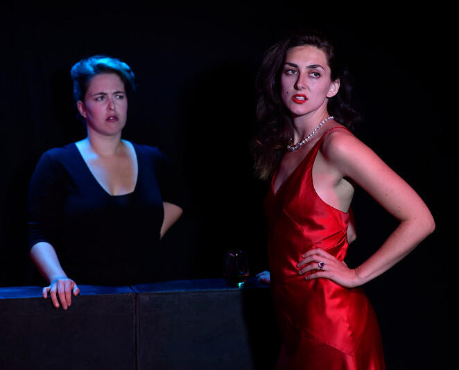 Erin on stage at Edinburgh Fringe. They are looking disgruntled at their colleague who has long brown hair and is in a red satin dress.