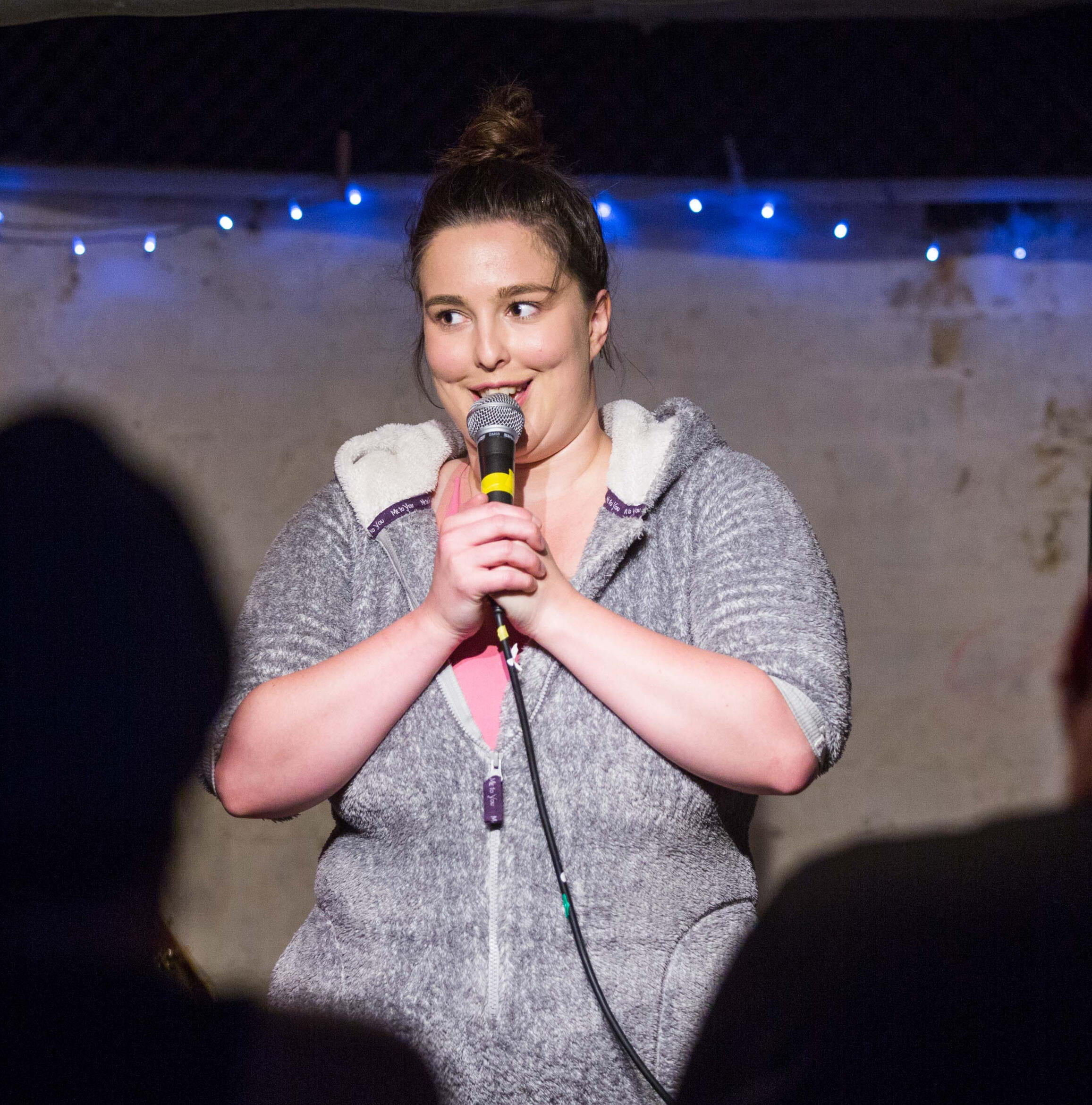 Erin on stage wearing a furry grey onesie. They are holding a microphone and smiling in front of two silhouetted audience members.