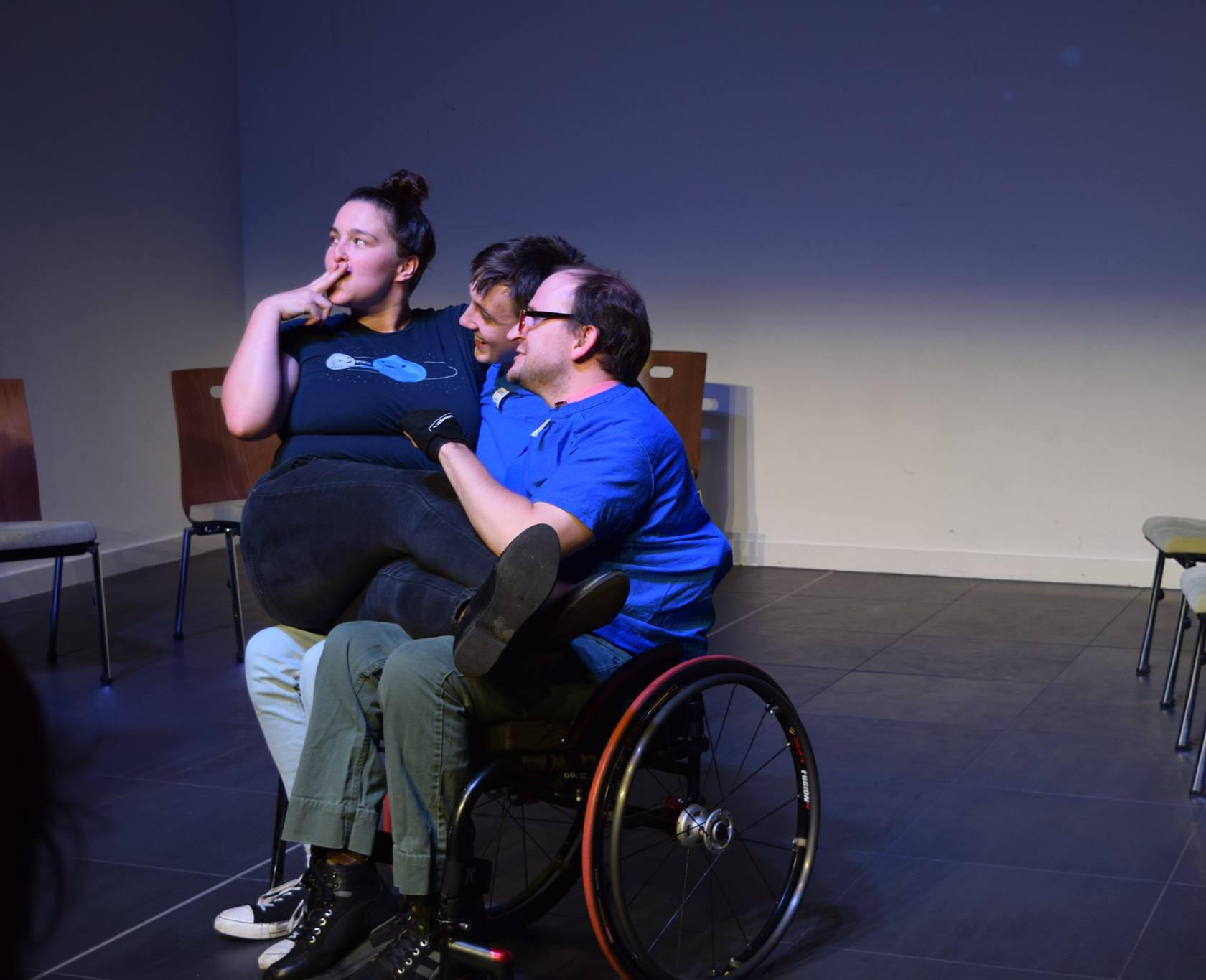 Erin sitting on the lap of a wheelchair-using colleague pretending to smoke a cigarette as part of an improv performance at The Waterside.