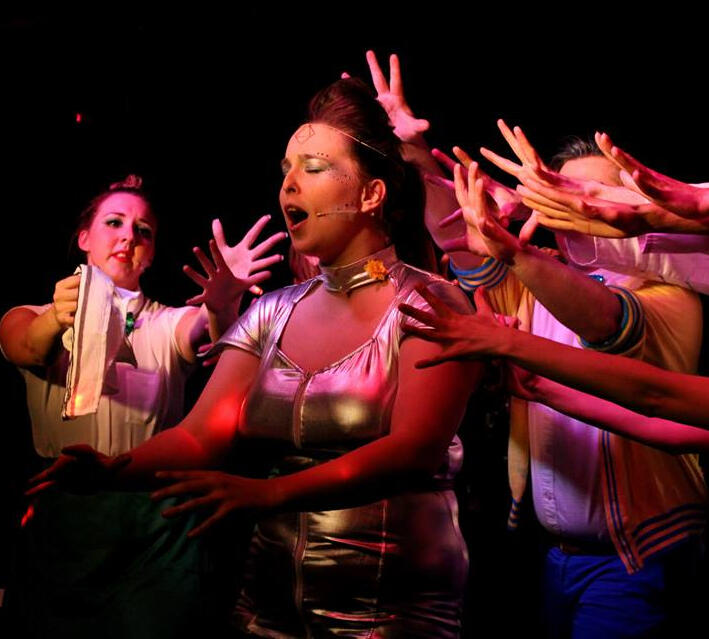 Erin performing in Saucy Jack and the Space Vixens. They are singing out to the audience and the ensemble's hands are reached out towards Erin.