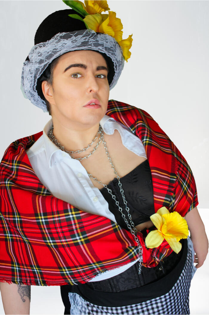 Erin in their genderbent welsh lady costume. They are wearing light drag king makeup and are looking up the camera fiercely. They are wearing a hat with daffodils on, a red tartan shawl, a chain necklace, a black bra and a white shirt.