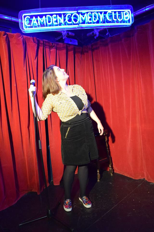 Erin on stage at the Camden Comedy Club looking up at the neon sign above their head.